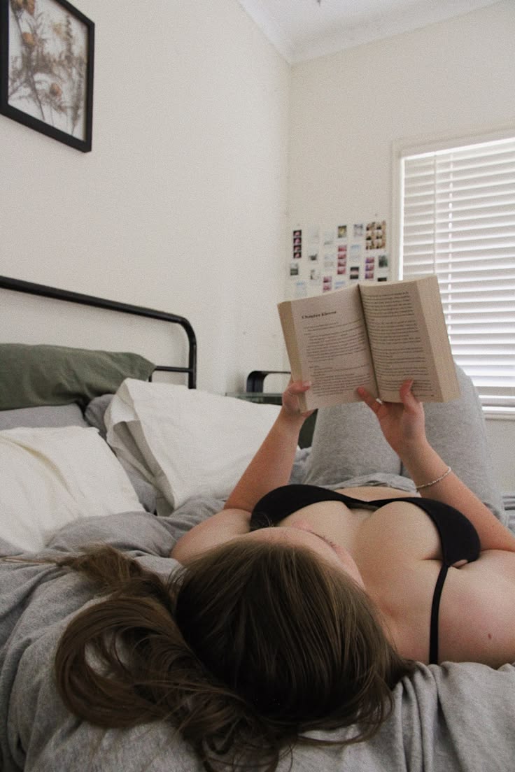 Person lying on a bed reading an open book in a bright bedroom.